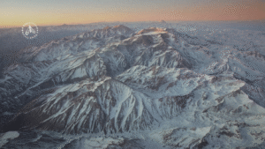 Vista aérea detalhada das montanhas andinas cobertas de neve com cristas escuras e sombreadas sob um céu rosa-laranja, destacando a geografia do medo e a estética do gótico andino.